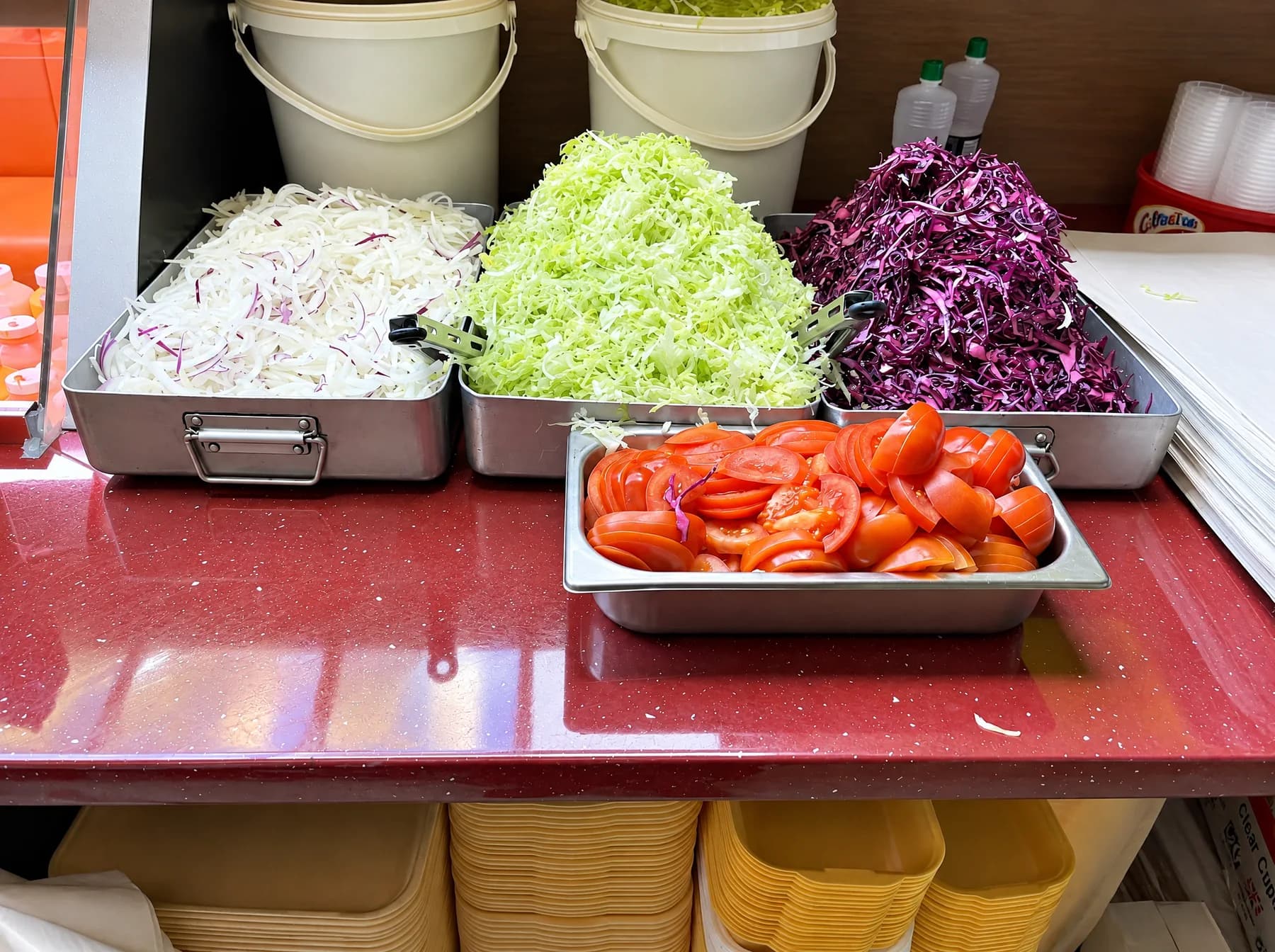 Steel trays of shredded white cabbage, lettuce, red cabbage and sliced tomato on the counter